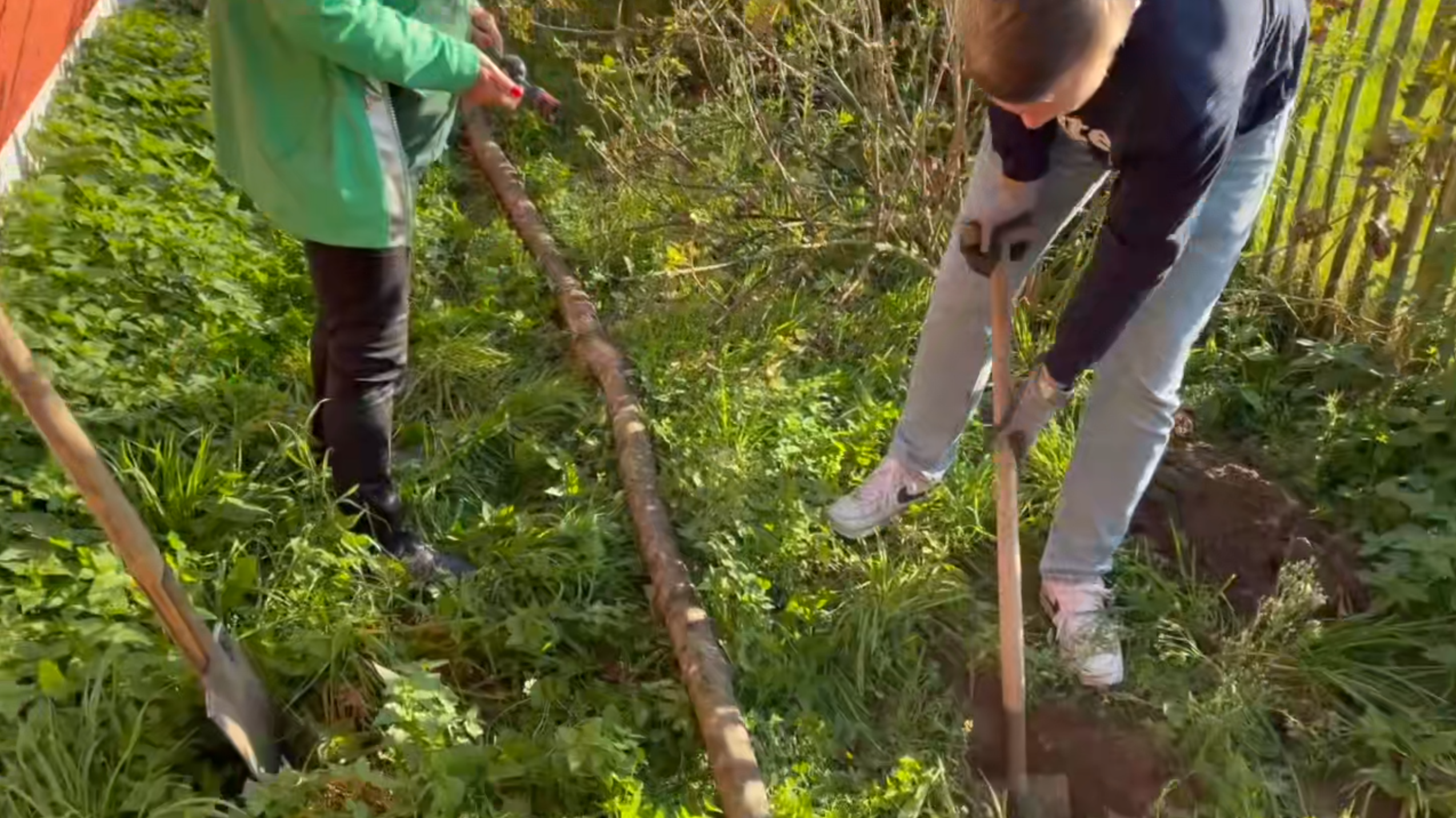 Max gräbt mit einem Spaten ein Loche in den Boden während Uschi einen Gartenschlauch anreicht.